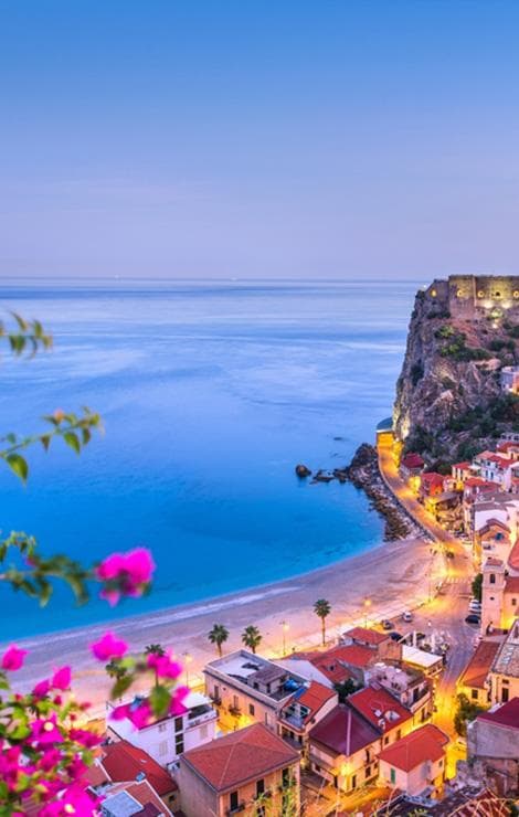 Panoramic view of a Sicilian coastal town at sunset, with a golden beach, colorful houses, and a cliff overlooking the sea.