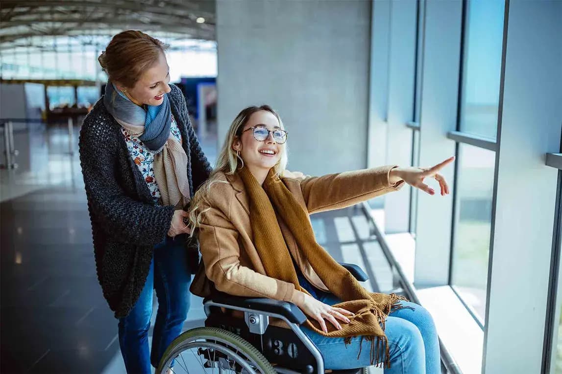 Donna su sedia a rotelle sorride e indica fuori dalla finestra in aeroporto, accompagnata da un'altra donna sorridente.